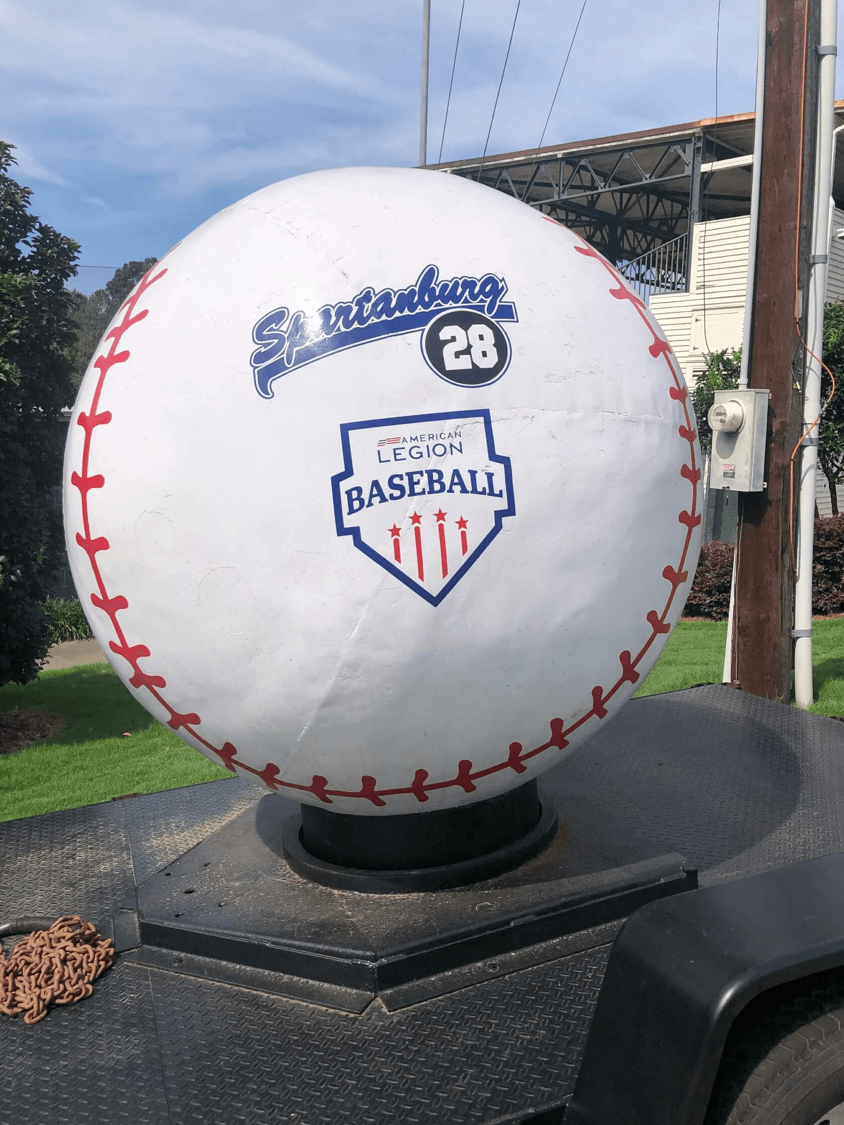 Giant baseball monument displaying the Spartanburg 28 script logo and the American Legion Baseball shield emblem, with the historic Duncan Park Stadium grandstand visible in the background.